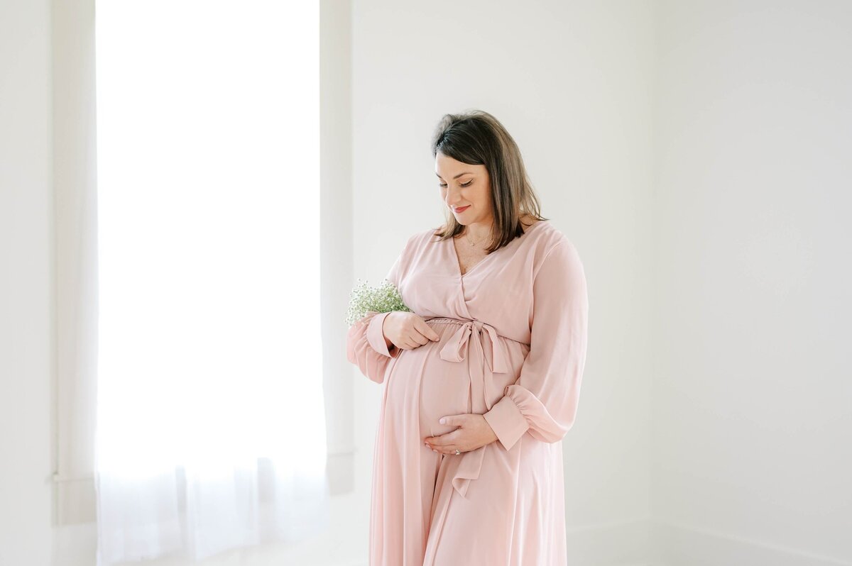 A pregnant brunette woman wearing a light pink dress cradles her bump with one hand and a bouquet of baby's breathe in the other.  Photograph captured  inside of Charlotte Maternity Photographer Melissa Mayrie Photography's  white, natural light studio.