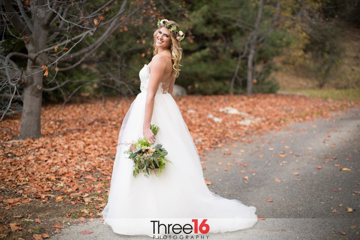 Bride looks back at the wedding photographer posing with her bouquet down by her side and dress train fanned out amongst the fall season leaves