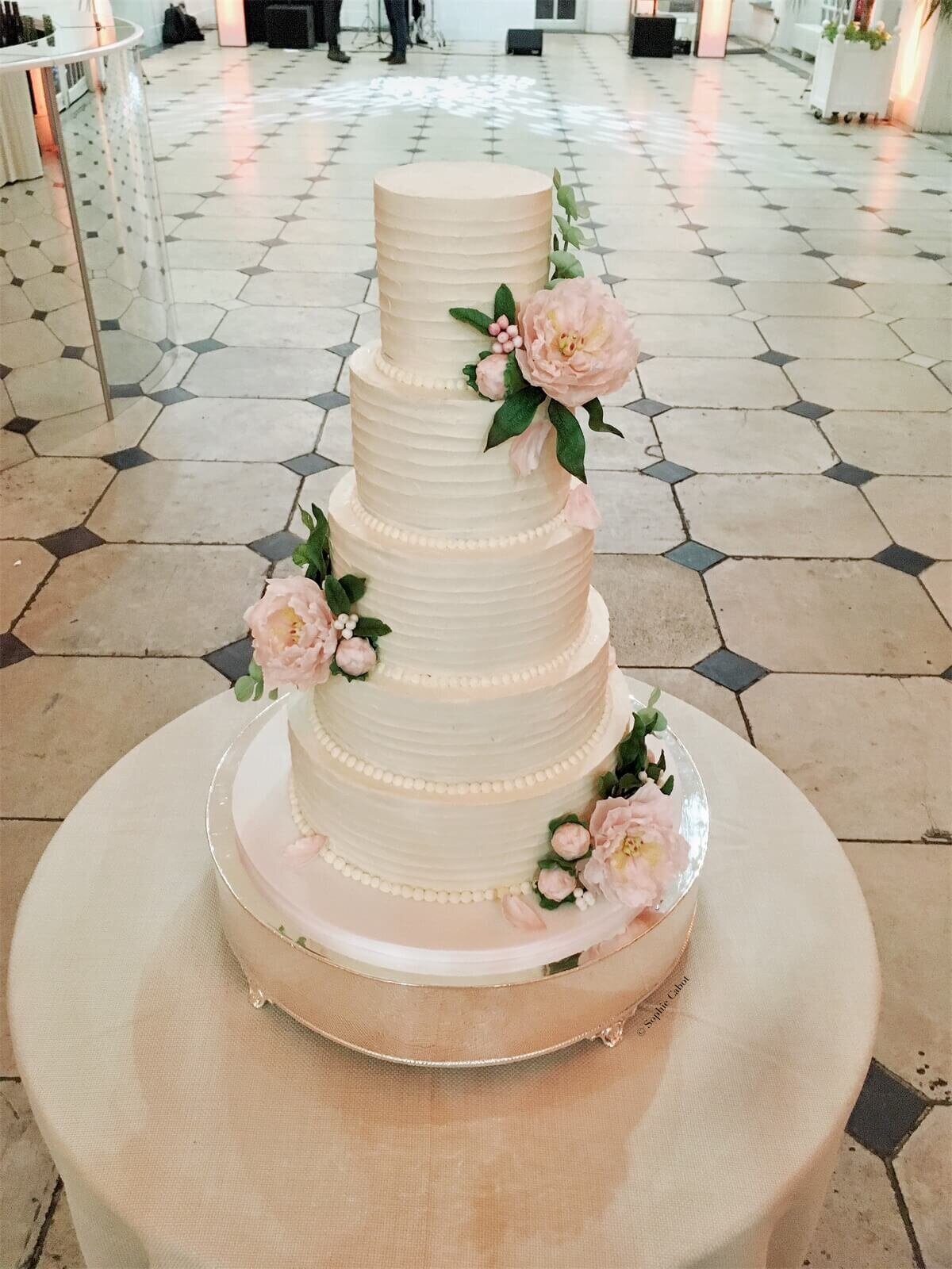 A white wedding cake with five tiers with a white ganache finish and sugar flowers clusters down the cake