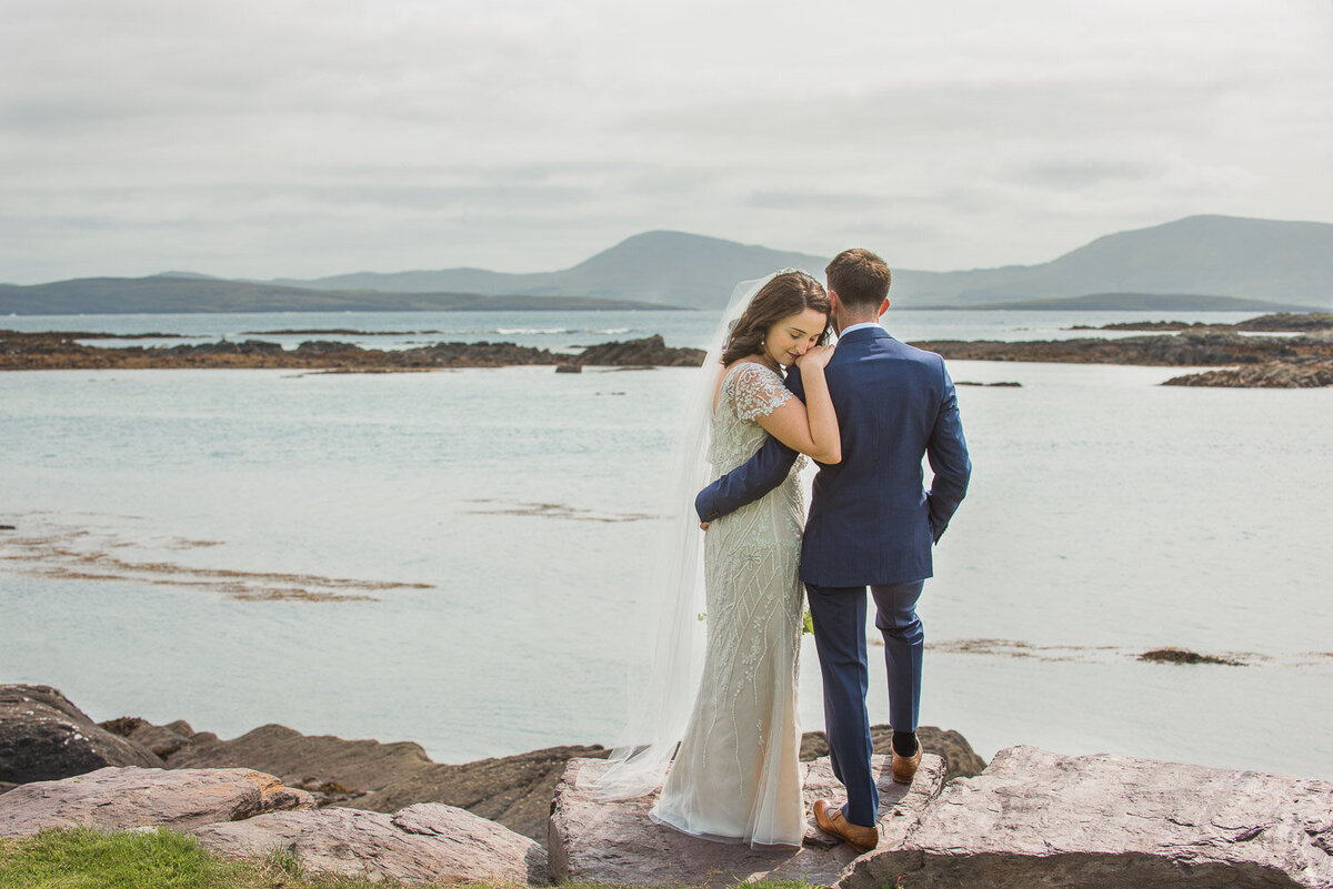 bride wearing a vintage beaded, column wedding dress hugging her husband during the first look while standing on a rock overlooking the beach in Kerry