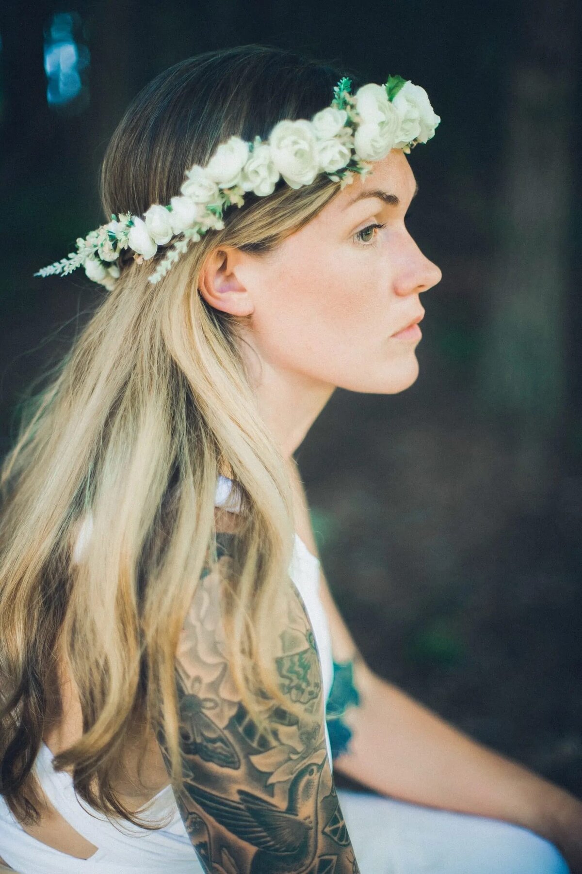 A contemplative woman wearing a floral crown, with her blonde hair cascading down her shoulder, set against a tranquil forest backdrop.