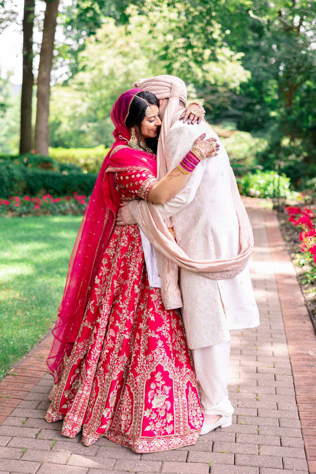 A couple embraces on a brick path in a garden. The woman wears a bright pink and white embroidered traditional dress with a matching headscarf and bangles, while the man wears a cream-colored traditional outfit. Trees and flowers surround them.