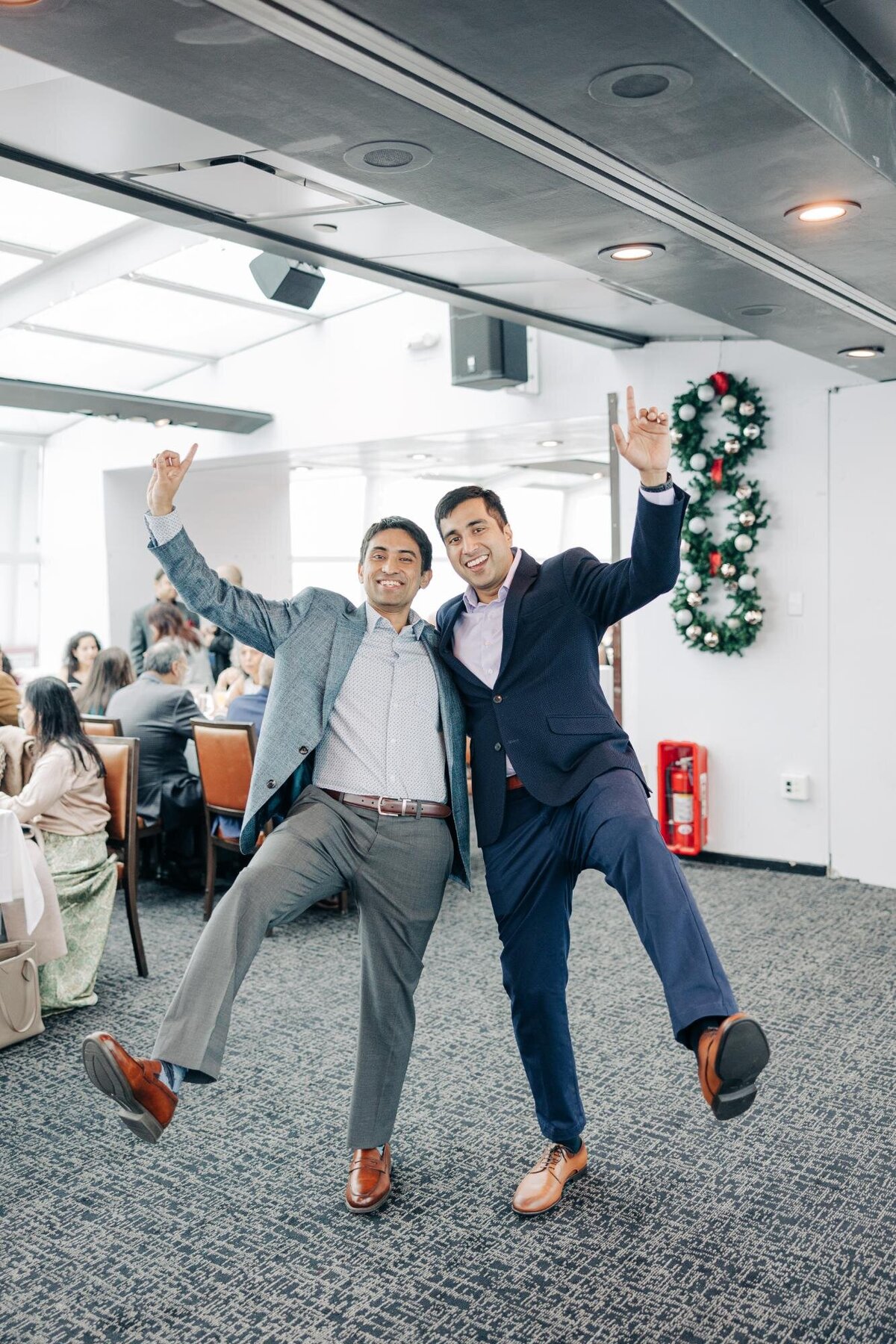 Two men in suits joyfully posing with one leg raised in a festive room with people dining in the background.