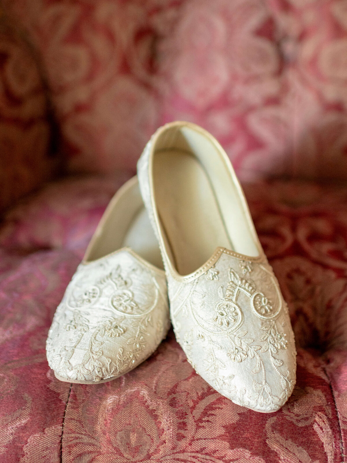A pair of elegant, off-white embroidered slip-on shoes rest on a plush, pink floral-patterned fabric surface. The intricate embroidery features floral designs, adding a touch of sophistication.