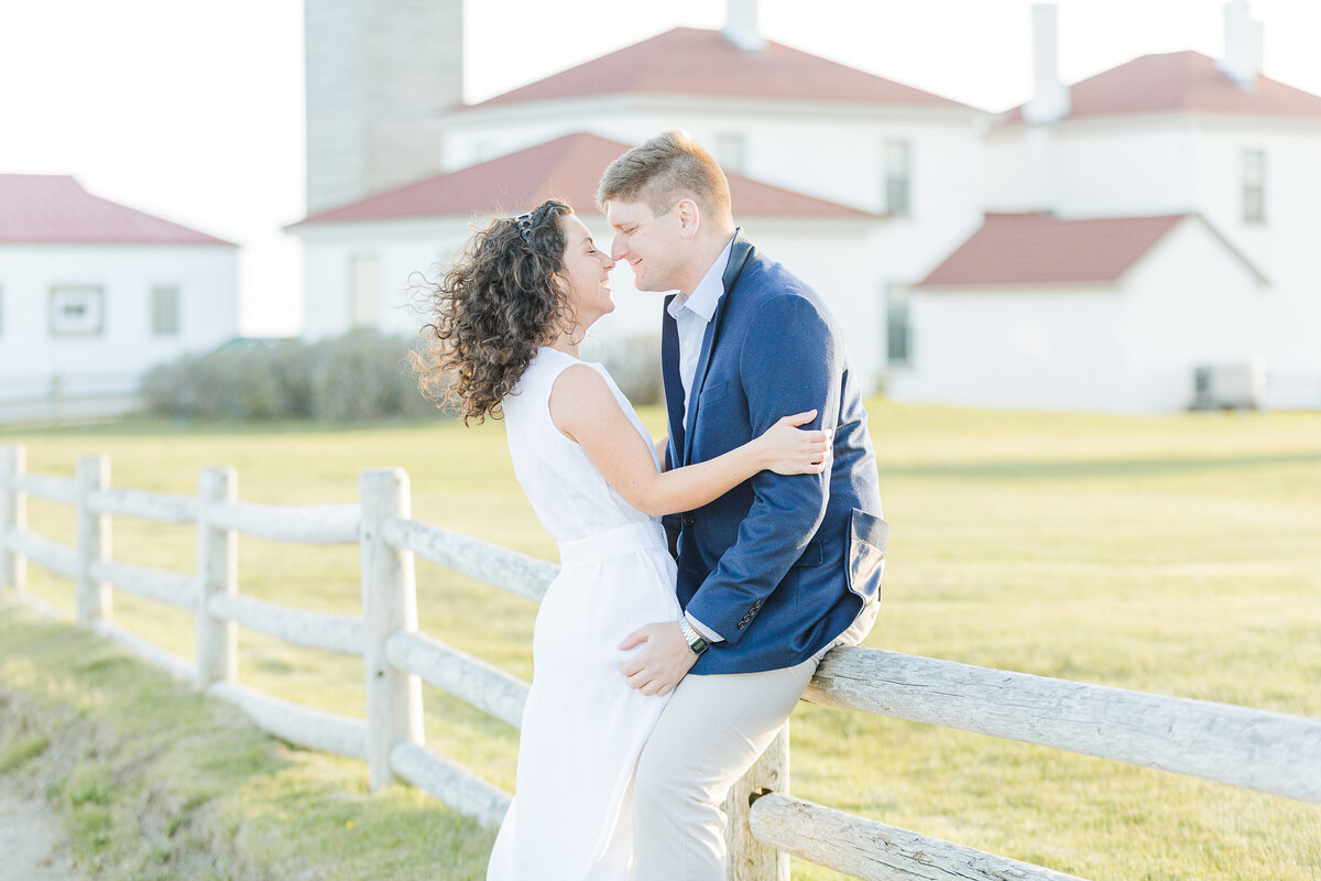A groom is leaning along the fence at Beavertail Lighthouse in Jamestown, RI for his engagement photoshoot. The woman is standing between his legs and their noses touching. The lighthouse is seen in the background. Captured by RI wedding photographer Lia Rose Weddings