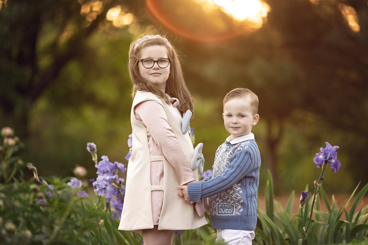 A young girl and her toddler brother hold hands among some purple flowers in a park at sunset