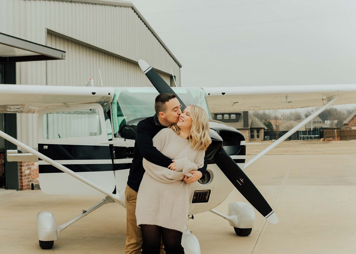 Maddie Rae Photography couple standing in front of an airplane. the guy is standing behind her and giving her a back hug and kissing her cheek. she is smiling back at him