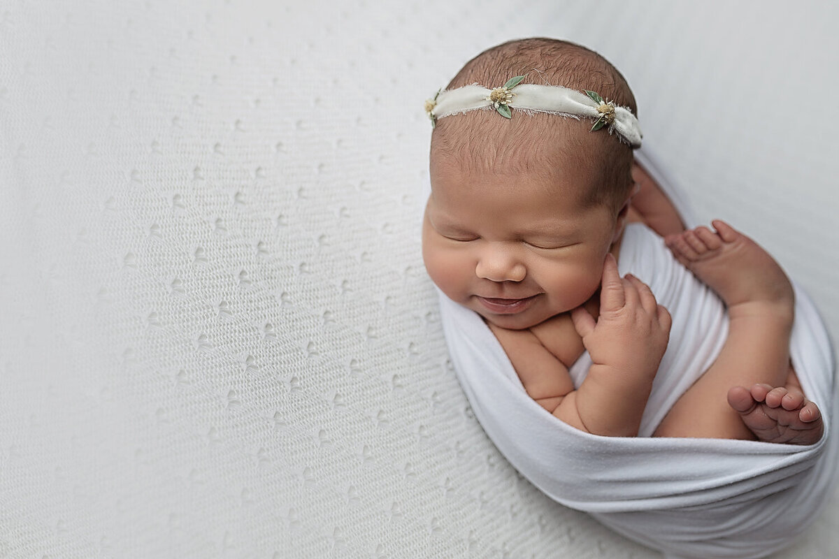 A newborn smiles while sleeping in an open white swaddle in studio