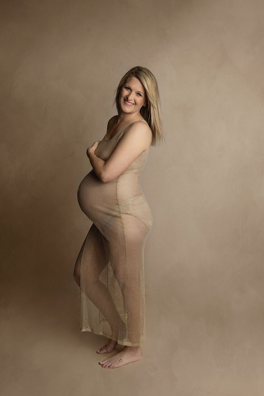 A mother to be stands barefoot in a studio in a sheer gold maternity gown covering with one arm