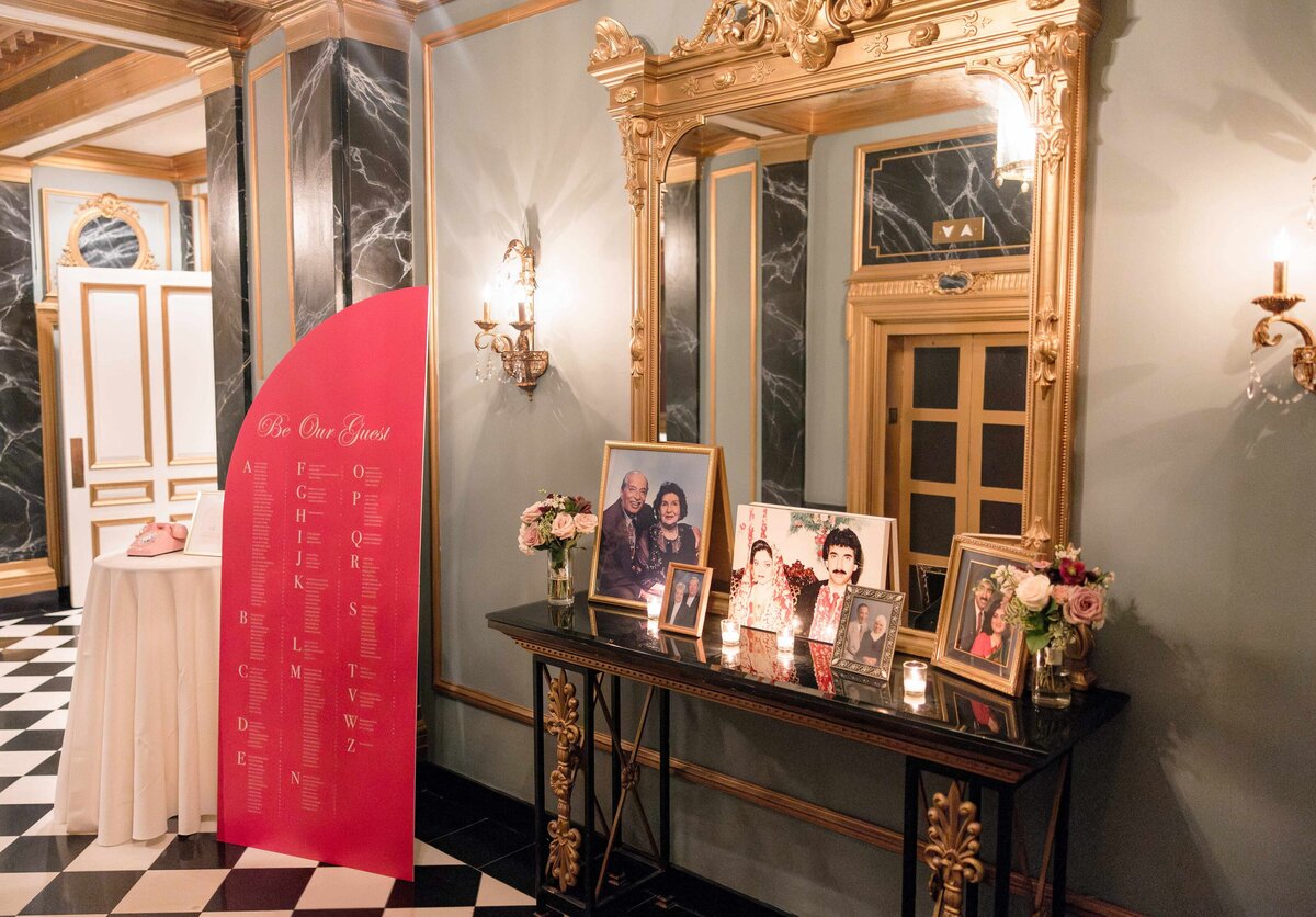 A luxurious hallway with a black and white checkered floor features a decorative table with family photos and candles. A pink sign with gold writing is placed nearby, surrounded by elegant gold-framed mirrors and sconces.