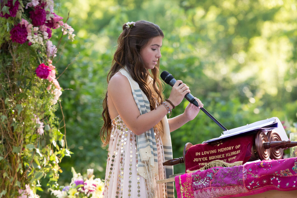 A teenage girl speaks into a microphone around pink flowers New York City bar mitzvah photographer Milestone Images