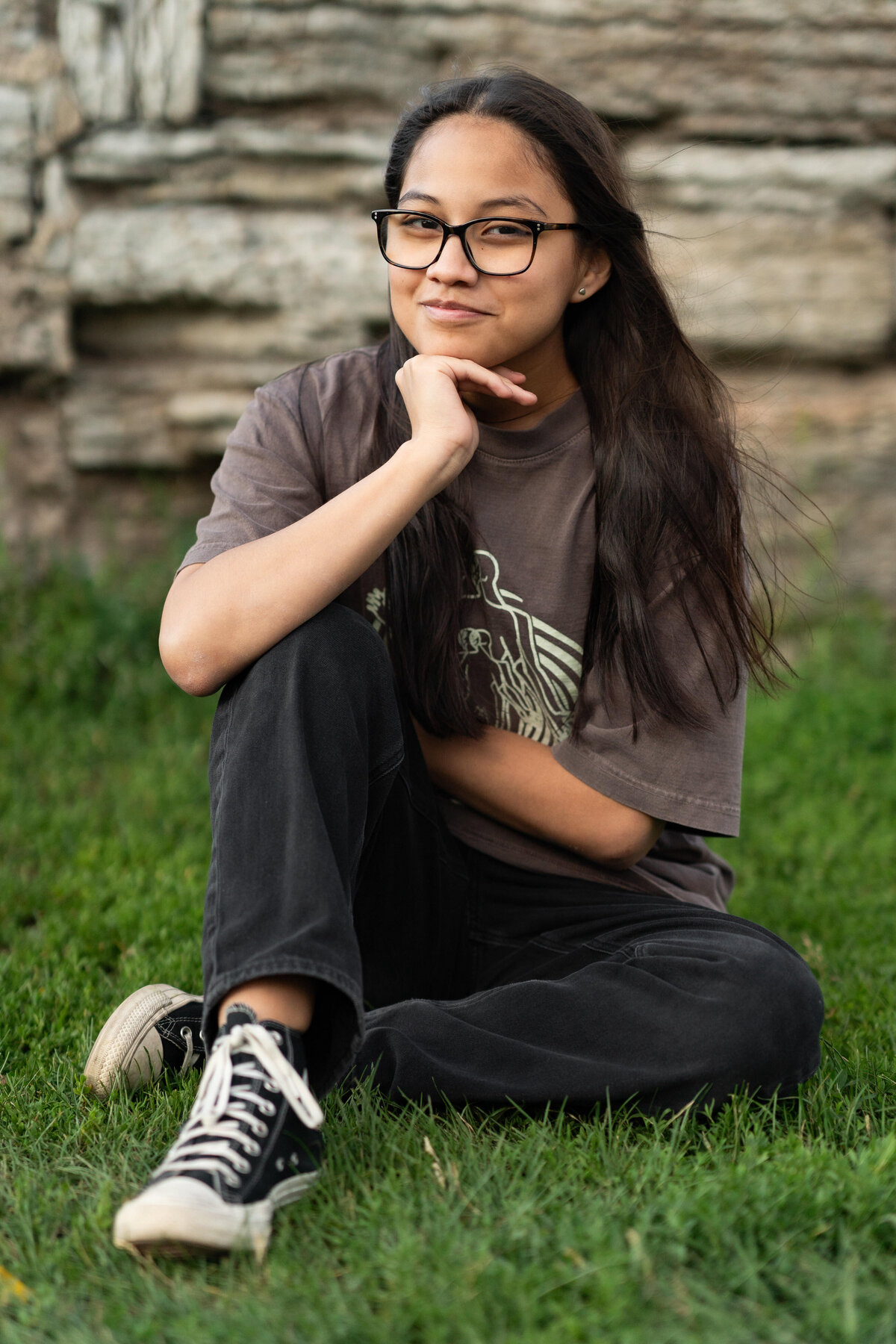 Senior girl in glasses and jeans smiles at the camera while sitting in the grass.
