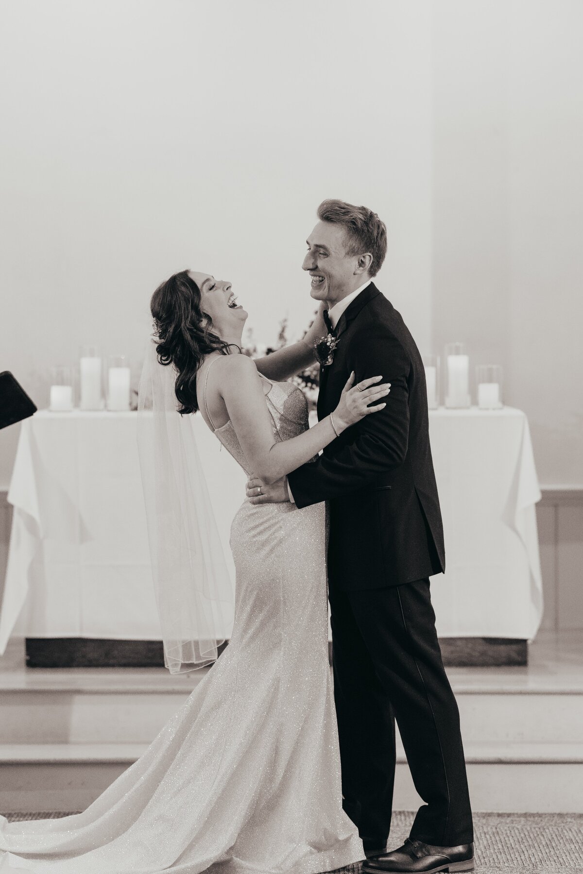 A joyful bride in a sparkling gown and a groom in a black suit dancing at their Iowa wedding reception, surrounded by candles and a draped table.
