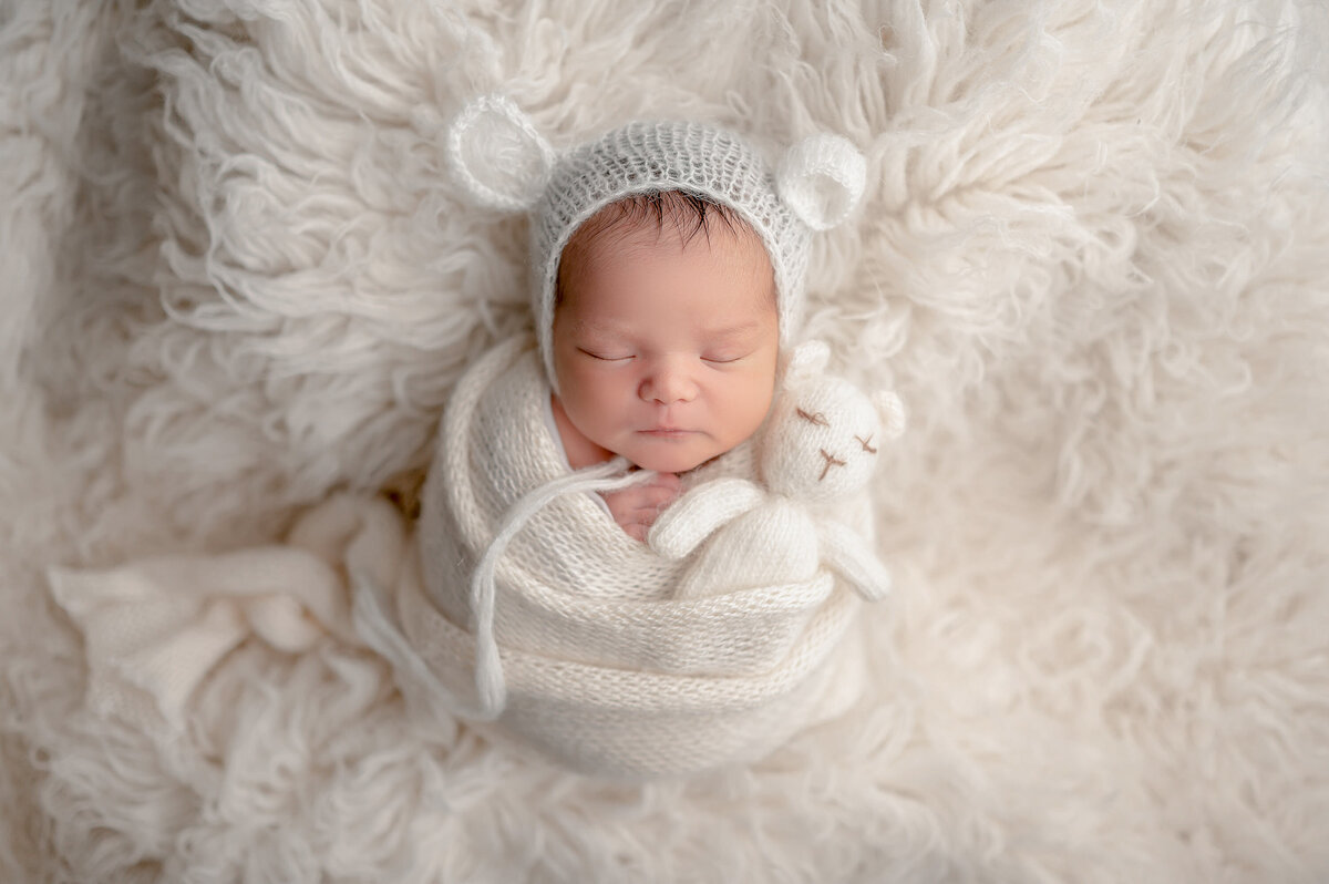 Newborn baby portrait showing infant wrapped in a woven, white blanket and matching hat laying on a furry, white rug.  Photographed in our Waukesha studio.