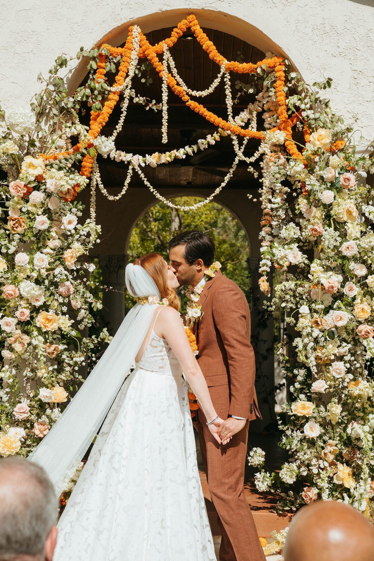 A couple kisses at their wedding ceremony underneath a large Anthousai floral installation in an arch.