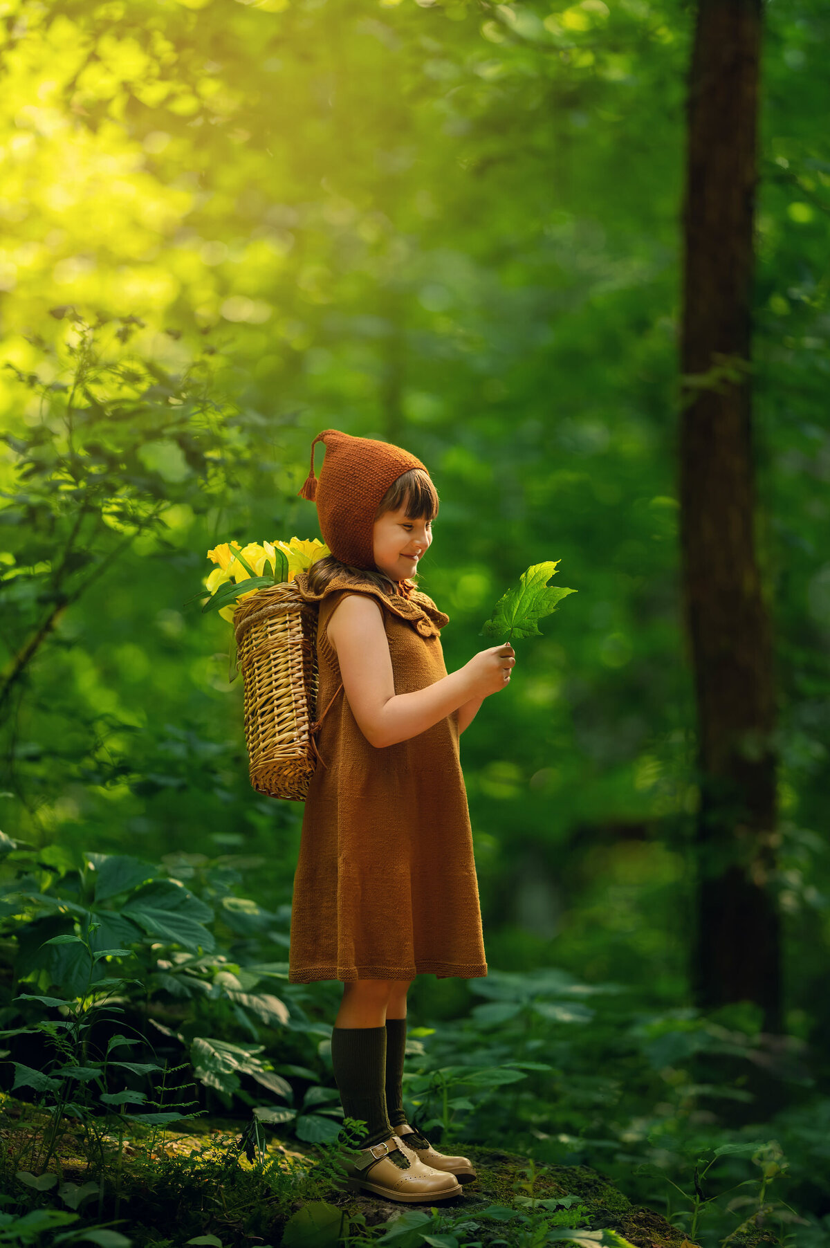 A young girl stands in a magical forest setting carrying a backpack of daffodils while being photographed by Kara Reese of Waukesha, WI.