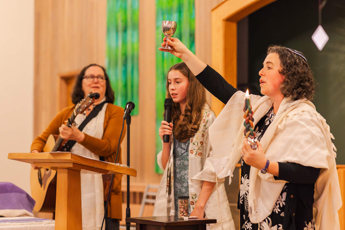 A rabbi lights a candle and holds a chalice on the bimoh with a guitar plays and teen girl singing