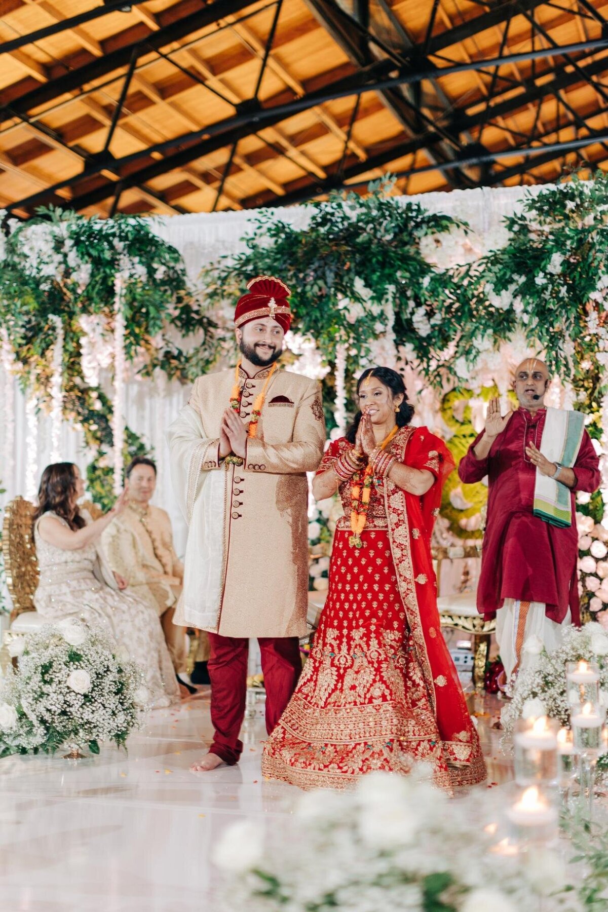 A bride and groom in traditional indian wedding attire participating in a ceremony surrounded by floral decorations and guests.