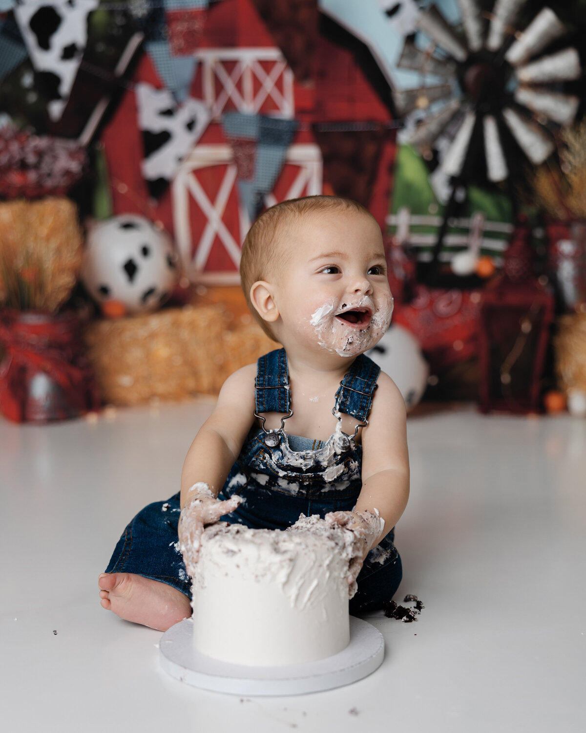 Cheerful one-year-old in blue overalls making a mess with his birthday cake in a farm-themed setup. Photograph by Yucaipa cake smash photographer Kaitlyn Dawn Photography.