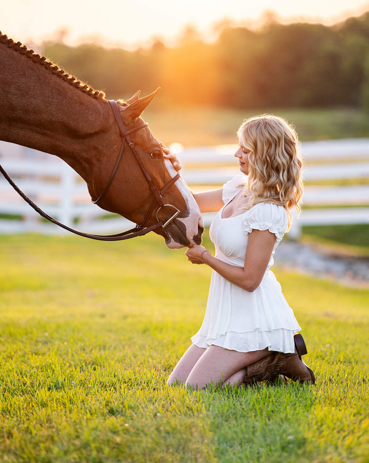 young blonde equestrian in a white jumper suit sitting on her knees in the grass with her chestnut paint horse nuzzled into her hands. behind them is beautiful white fence, trees, and golden hour sun.