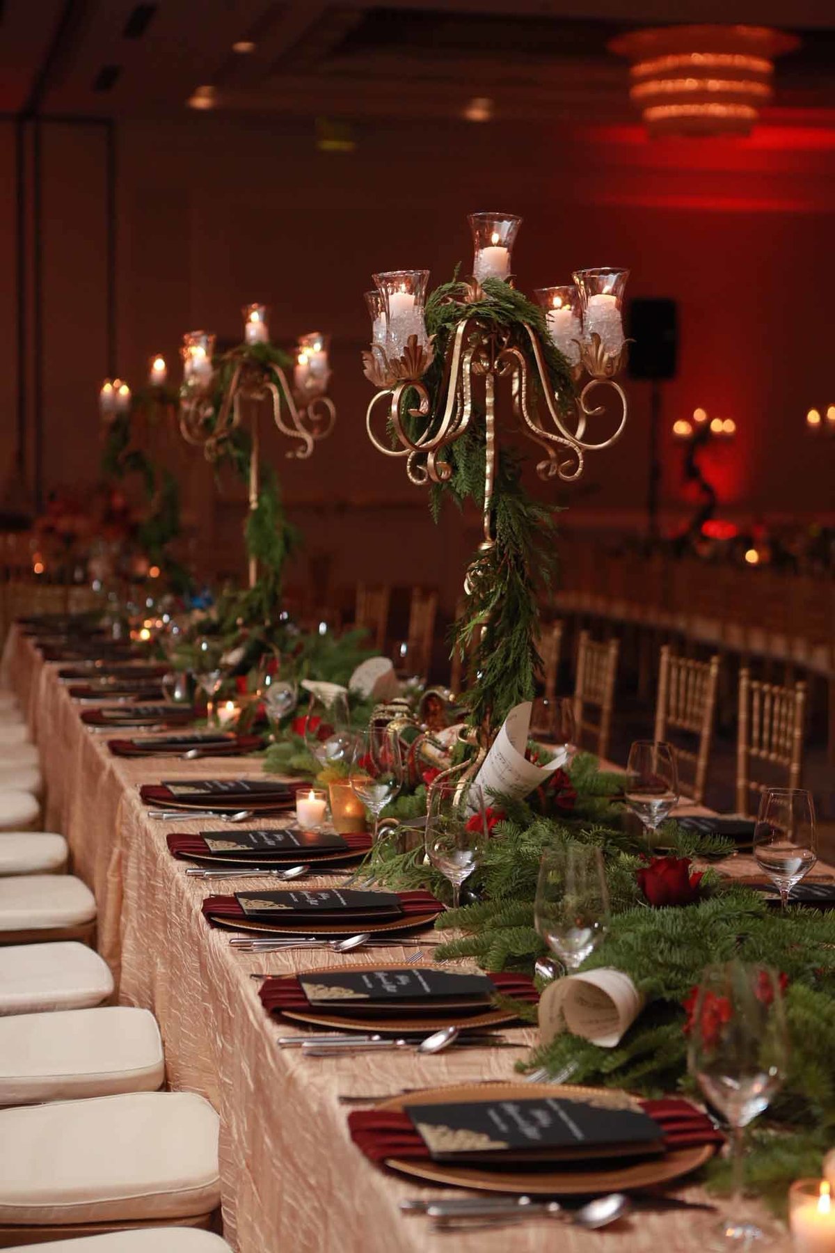 long table of tall gold candelabras, evergreen garland with red roses, gold charger plates, burgundy napkins for corporate holiday party