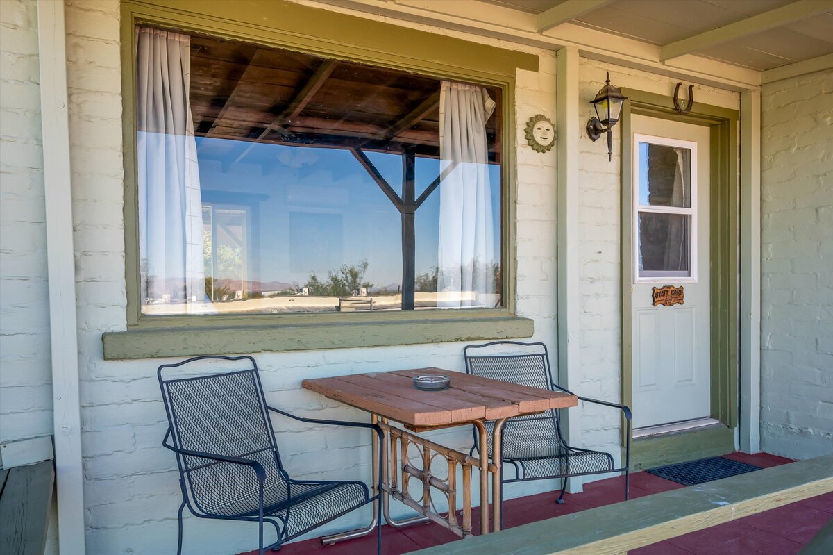 A small porch with a wooden table and two metal chairs in front of a white brick building with green trim.