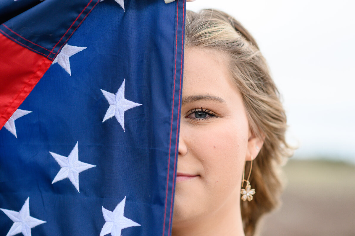 A denver senior girl hides half of her face with an american flag while looking at a Denver senior photographer