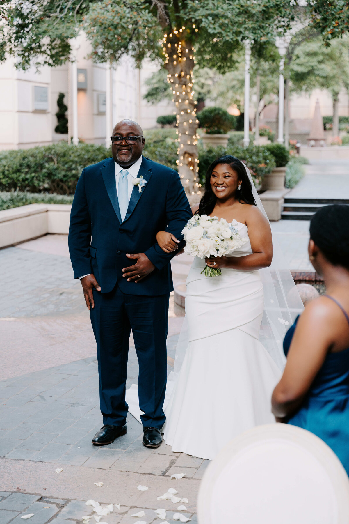 A bride in a white gown holds a bouquet, walking arm-in-arm with a man in a blue suit down an outdoor path adorned with lights, while guests watch.