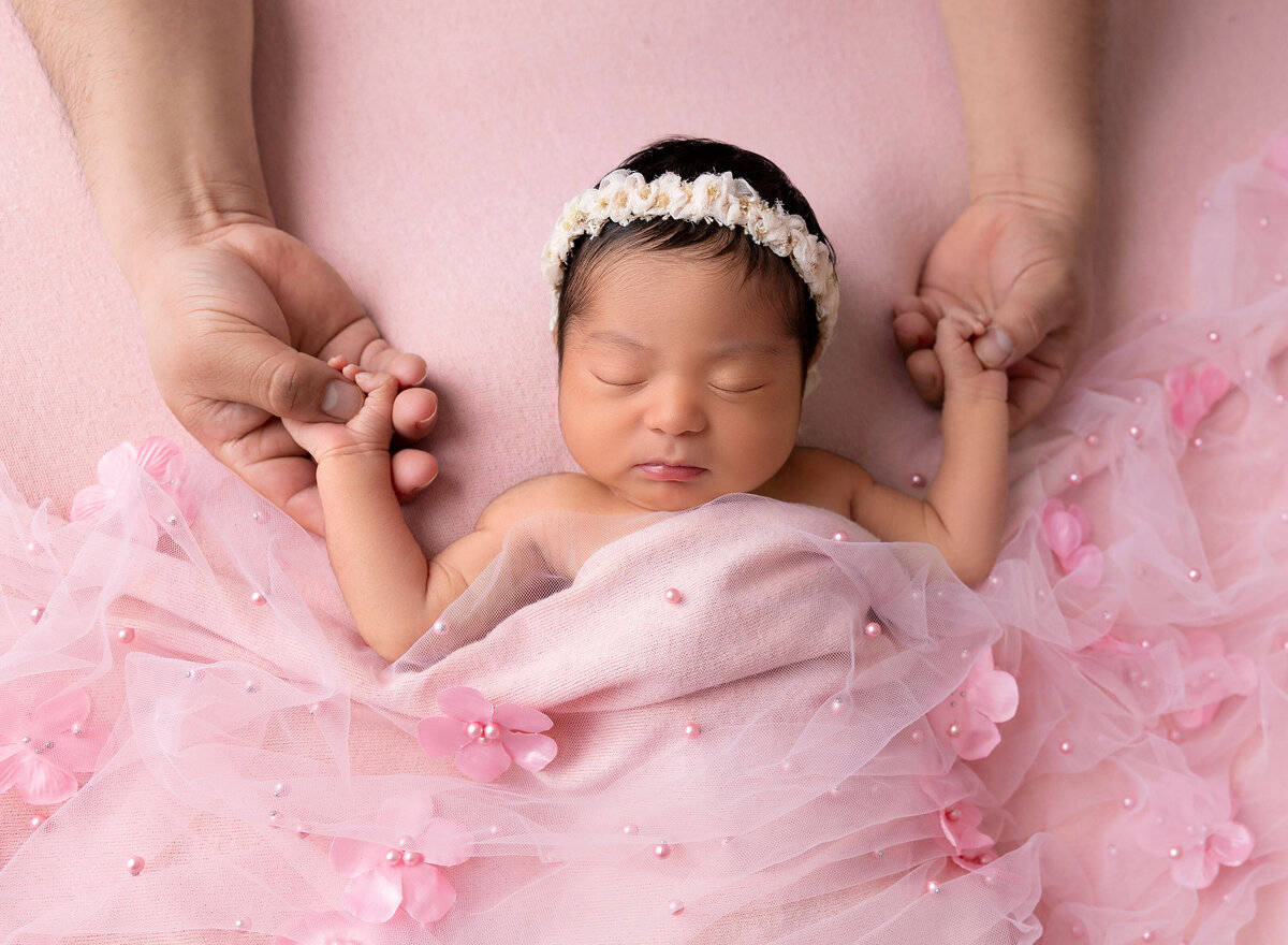 Aerial image from Brooklyn NY newborn photoshoot. Newborn baby girl sleeping on her back and her body is draped in a pink wrap embellished with pink pearls and flowers. You see only the dad's forearms. Baby girl is holding her dad's thumbs.