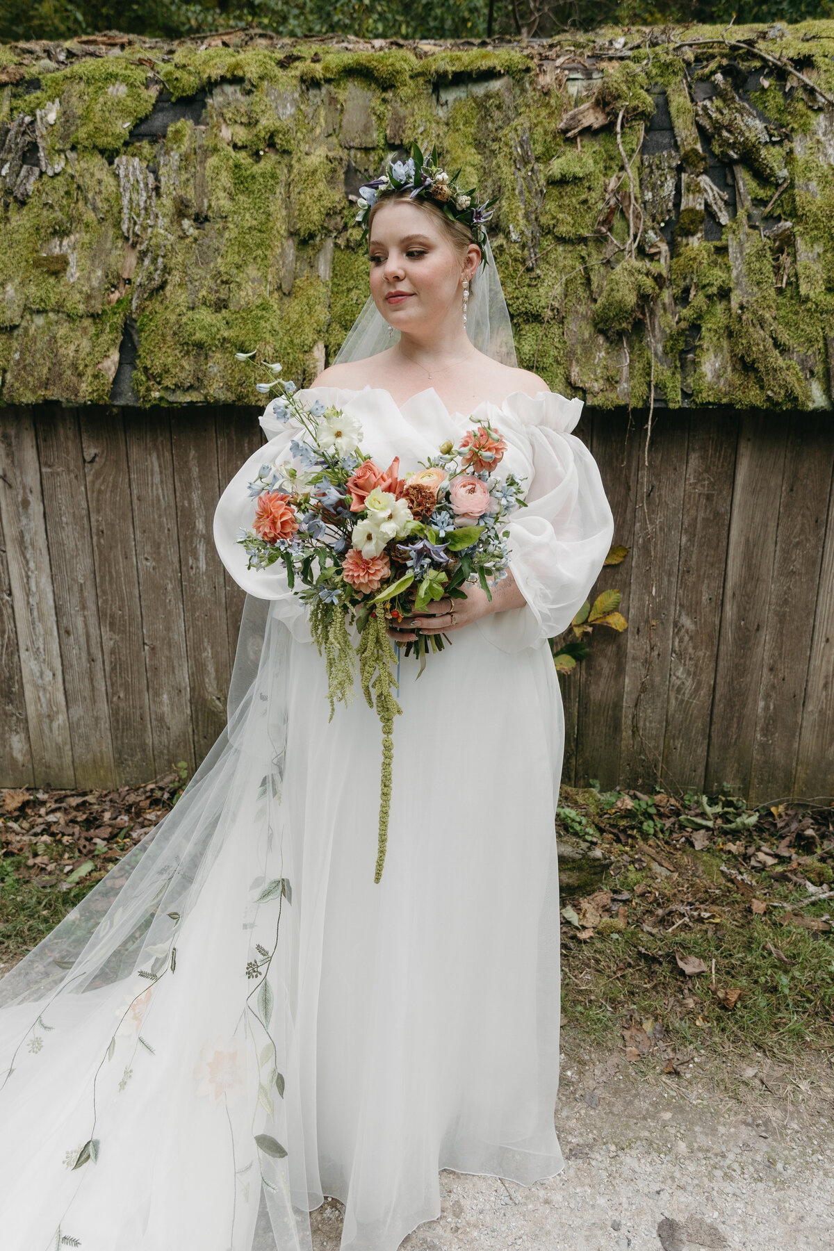 Bride holding a vibrant garden-style bouquet featuring dahlias, roses, delphinium, and greenery, wearing an off-the-shoulder tulle gown with embroidered veil, photographed against a rustic moss-covered wall.
