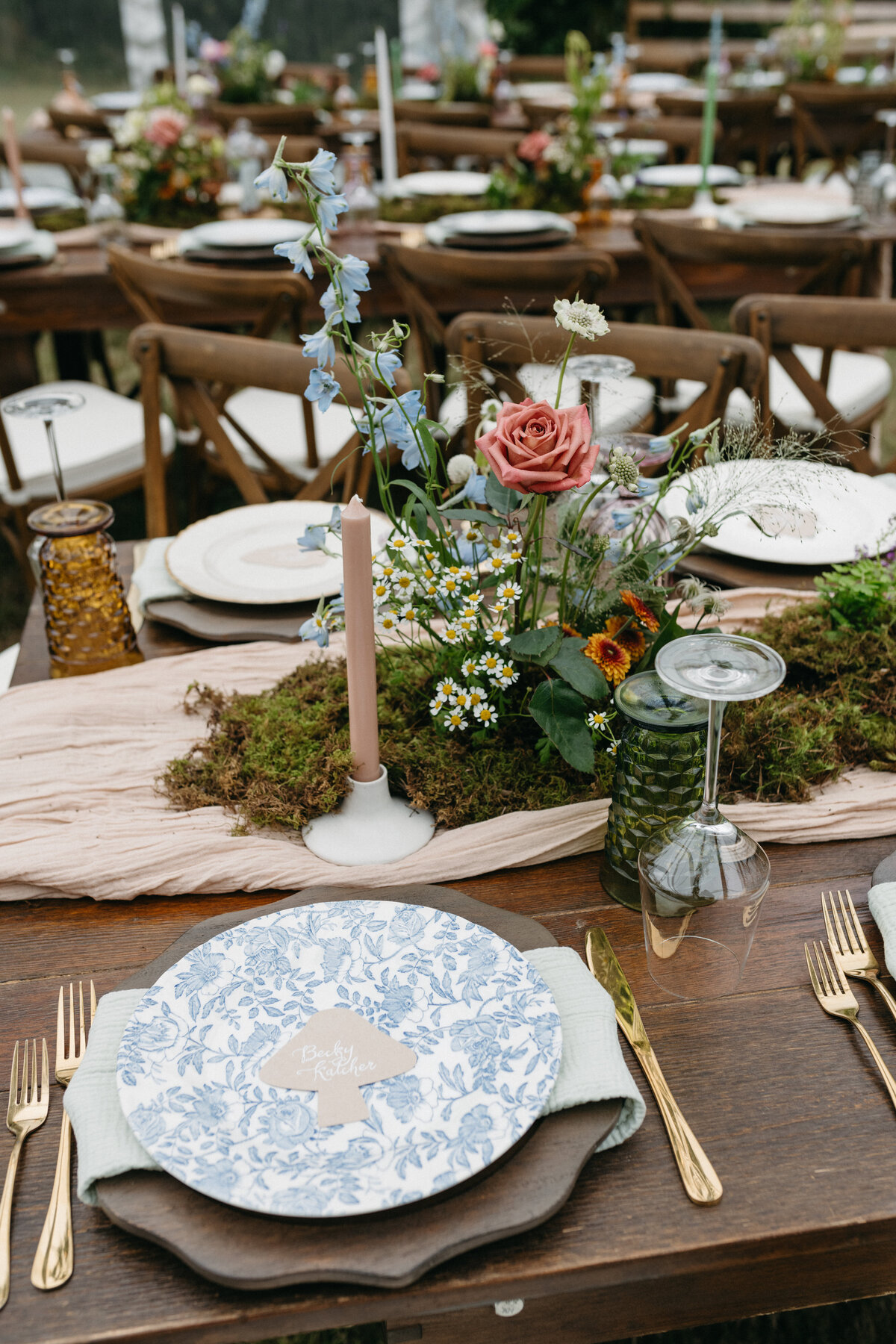 Romantic garden-inspired wedding reception table setting with blue floral china, gold flatware, textured linens, blush taper candles, and a whimsical floral centerpiece featuring roses, delphinium, chamomile, and moss, created by an Arkansas wedding florist for an outdoor tented celebration.