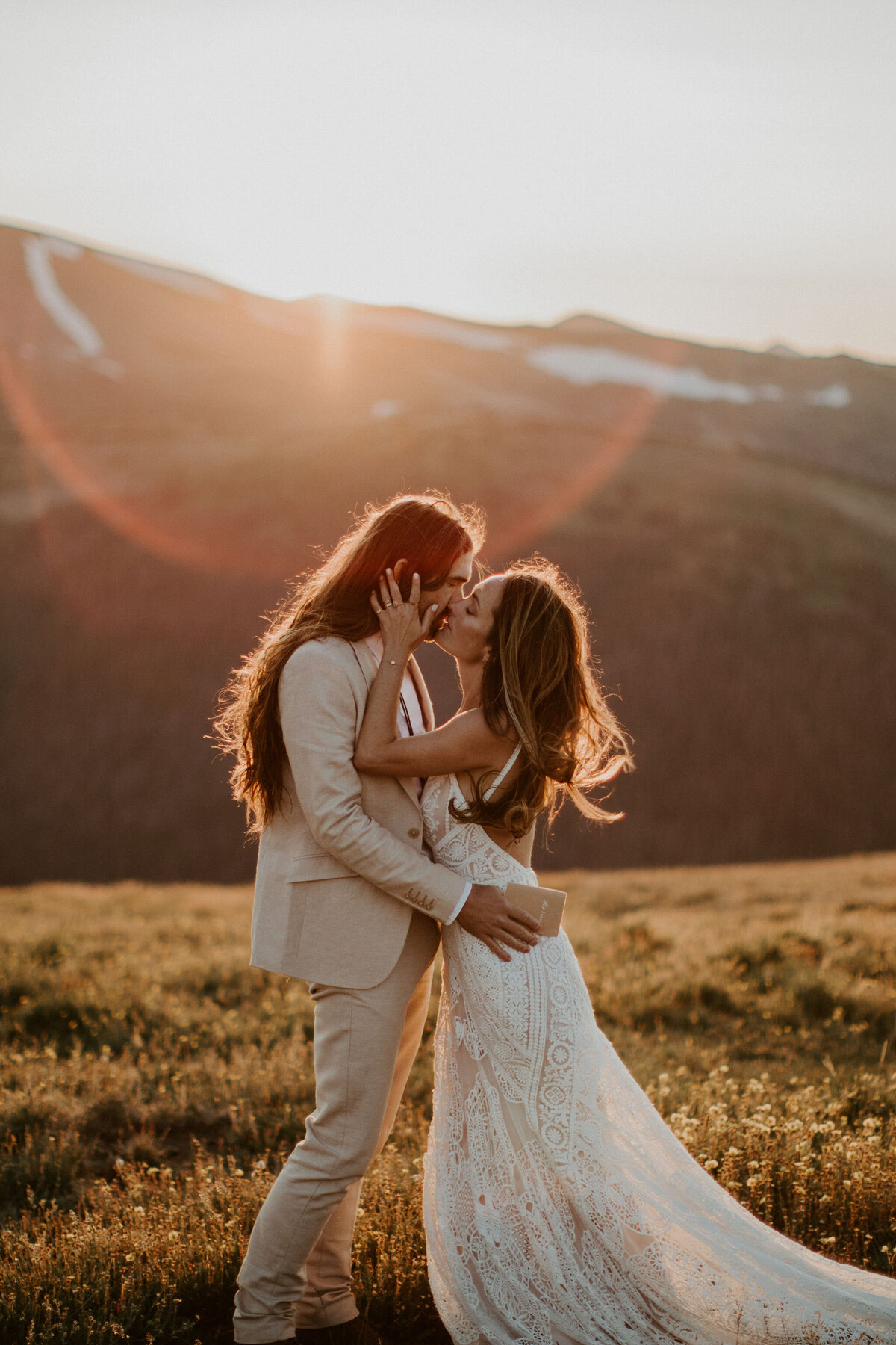 Bride and groom wearing an ivory suit and white wedding gown, kissing during golden hour holding tan wedding stationery.