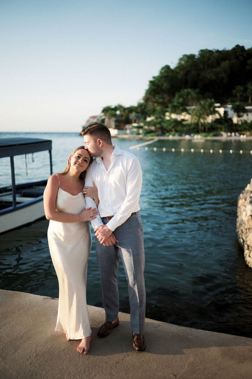 The engaged couple is standing on a dock, while the man is kissing the woman's head, at Round Hill Hotel & Villas, Jamaica.