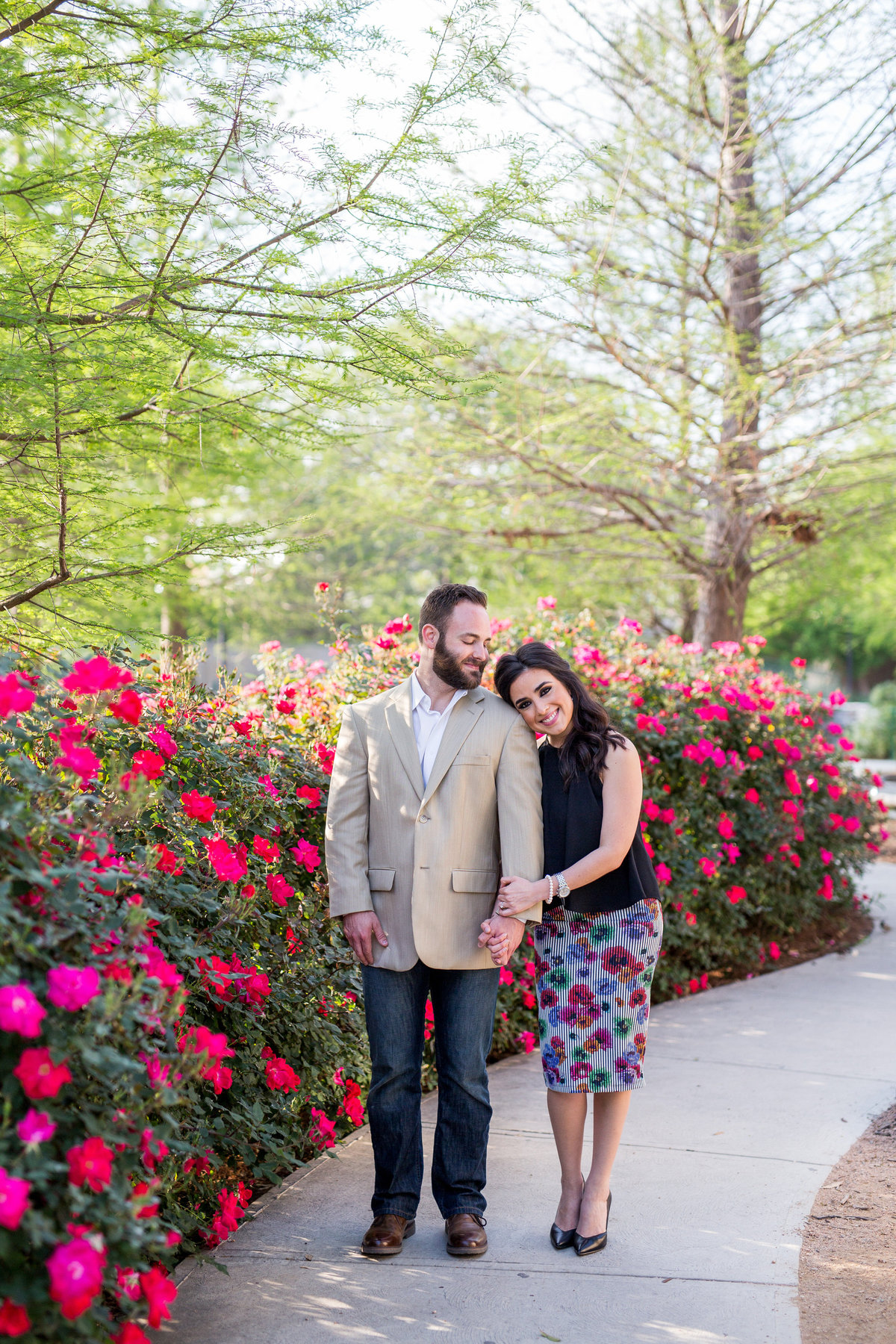 Engagement photography session of woman putting her head on the shoulder of her fiancé standing next to roses and the San Antonio Riverwalk.