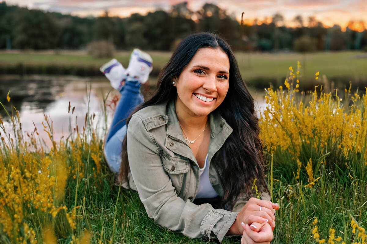 A gorgeous brunette is laying on her belly in a field of yellow wild flowers with a [pond behind her at sunset for her senior photo session.