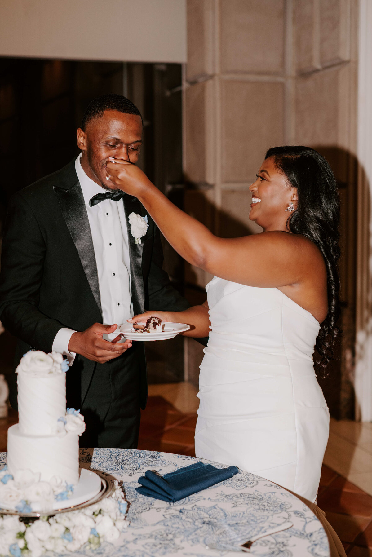 Bride in white dress feeds groom cake; both smiling, standing by a decorated white cake. Joyful wedding moment captured indoors.