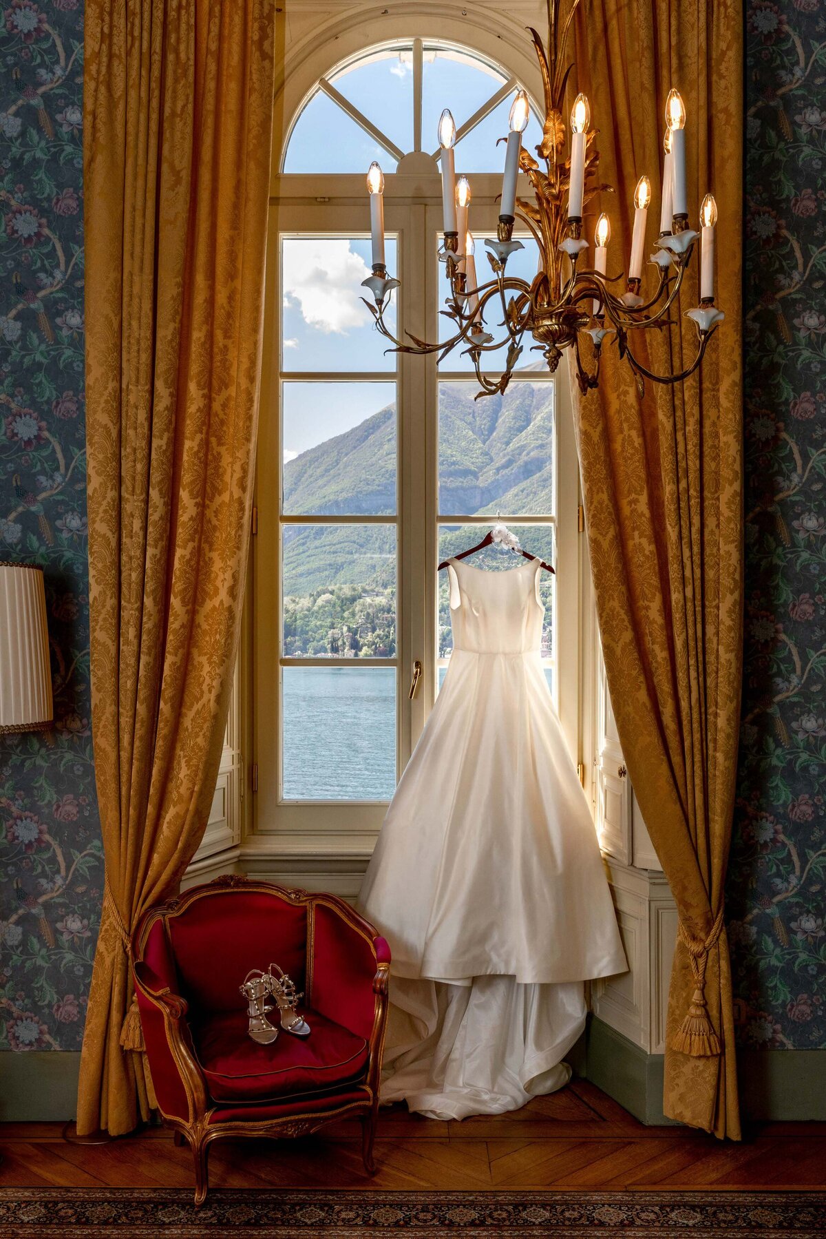 A wedding dress hanging up in a window with shoes on the chair next to it.