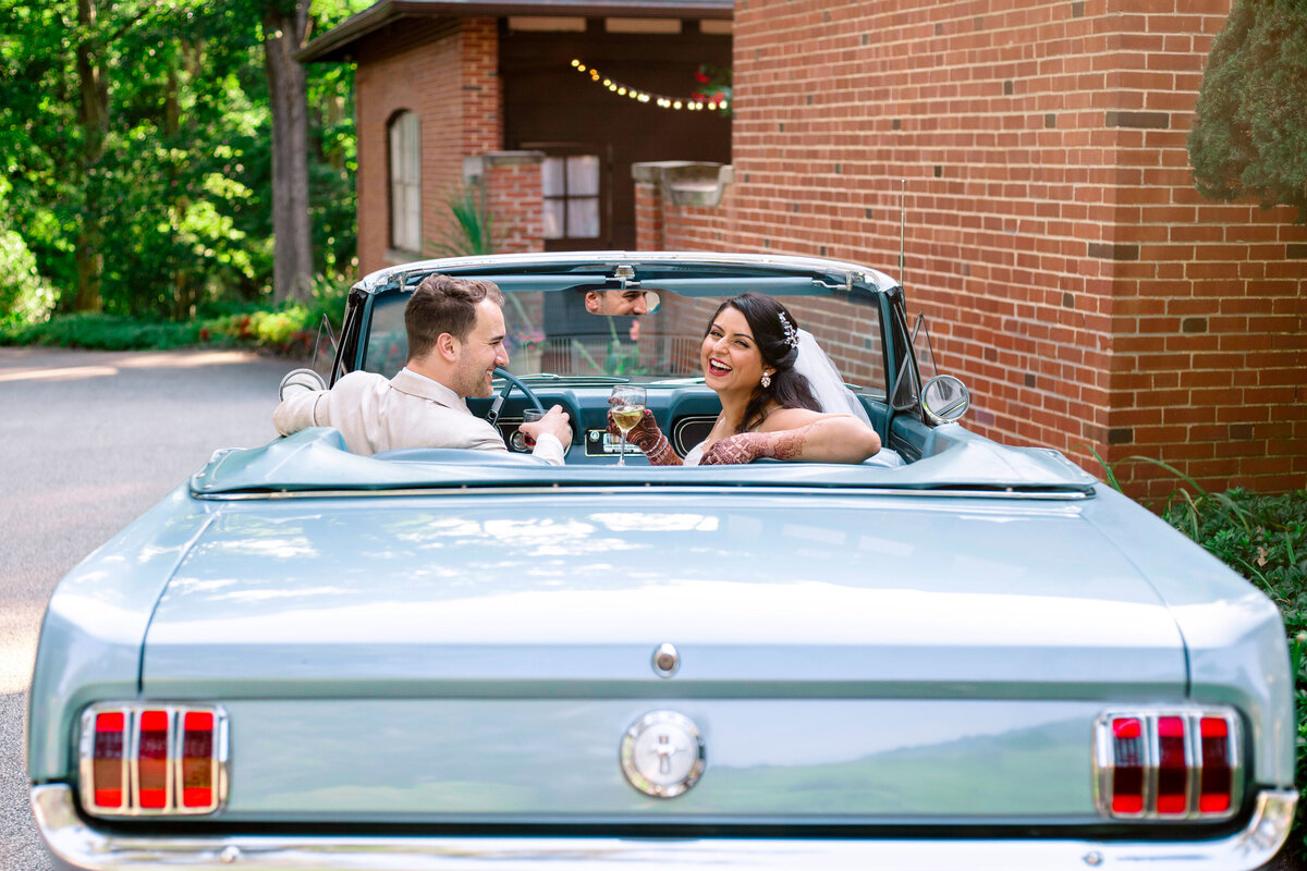 A newlywed couple sits in a vintage light blue convertible, smiling at each other. The groom, wearing a light-colored suit, drives while the bride, in a white dress and veil, holds a bouquet. The car is parked near a brick building with greenery around.
