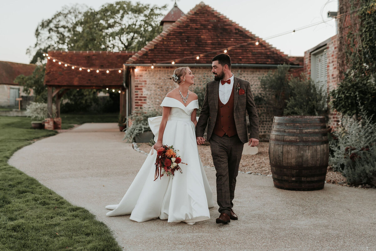 Bride & Groom walk hand in hand along a cobbled pathway at their rustic Autumn wedding at Montague Farm, Hankham, Sussex