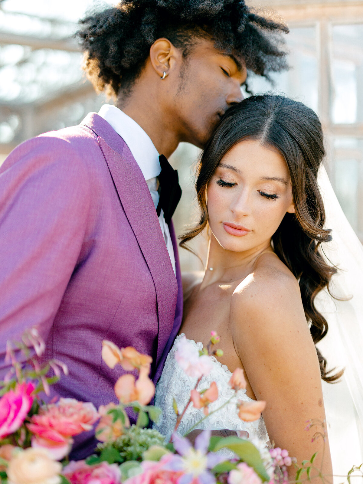 Groom in purple suit kissing bride on the head while she looks down