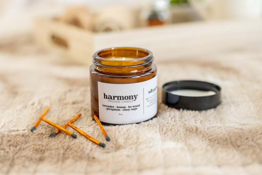 A jar of "harmony" candle on a textured fabric with three orange sticks beside it. the jar lid is off, showing the golden-wax candle inside.