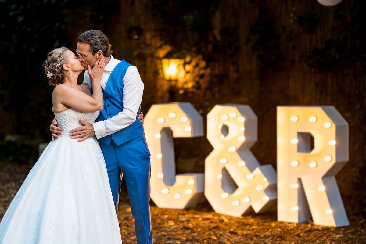 Bruidspaar poseert in de avond voor lichtletters met hun initialen, perfect georganiseerd door weddingplanner