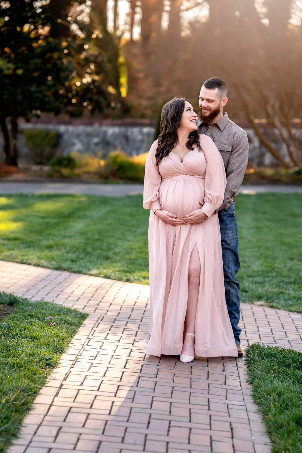 A mama to be in a long pink dress cradles her bump and smiles at her husband behind her