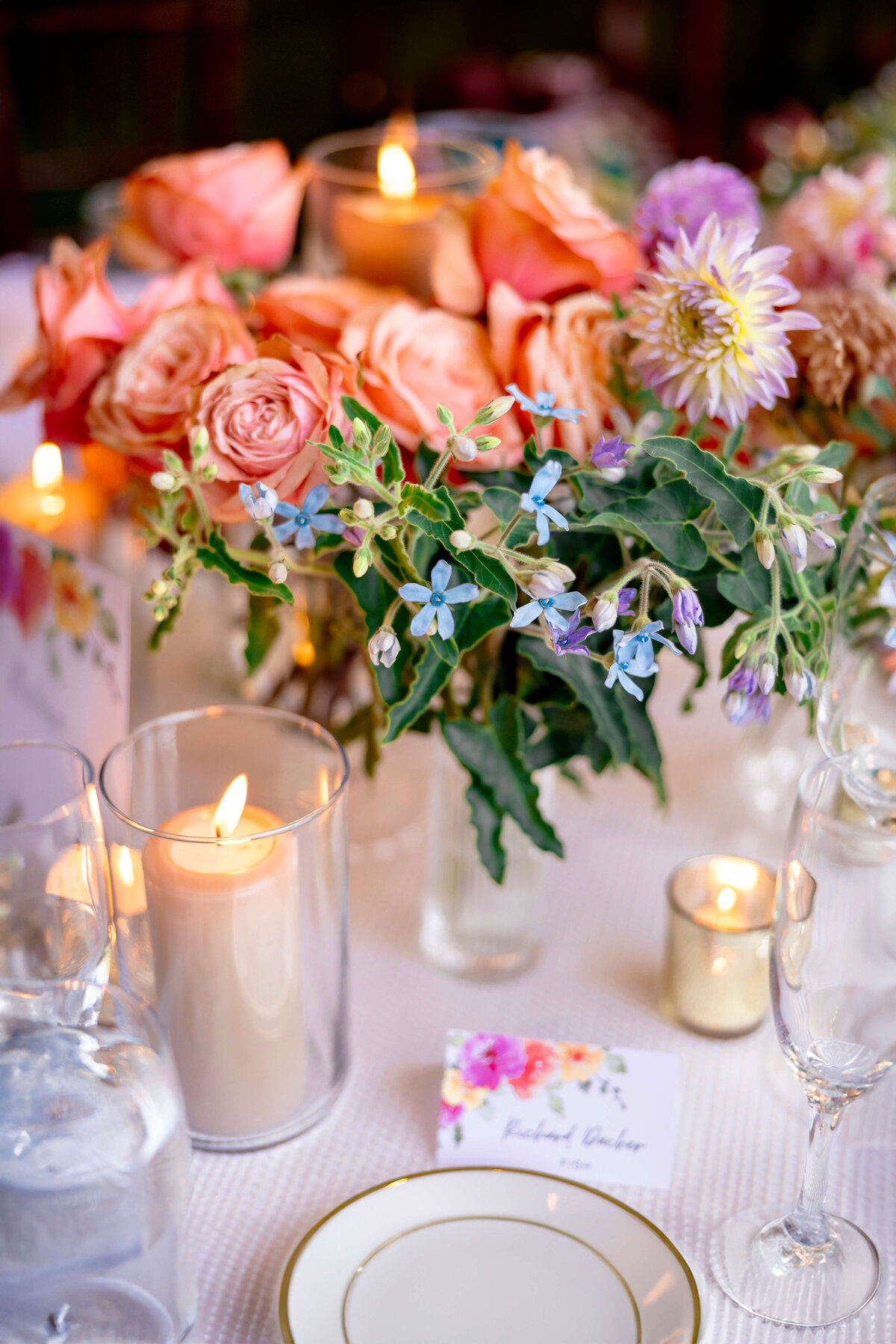 A beautifully set table with lit candles and floral centerpieces featuring pink and orange roses, along with small blue flowers. A place card and wine glasses are visible in the soft, ambient candlelight.