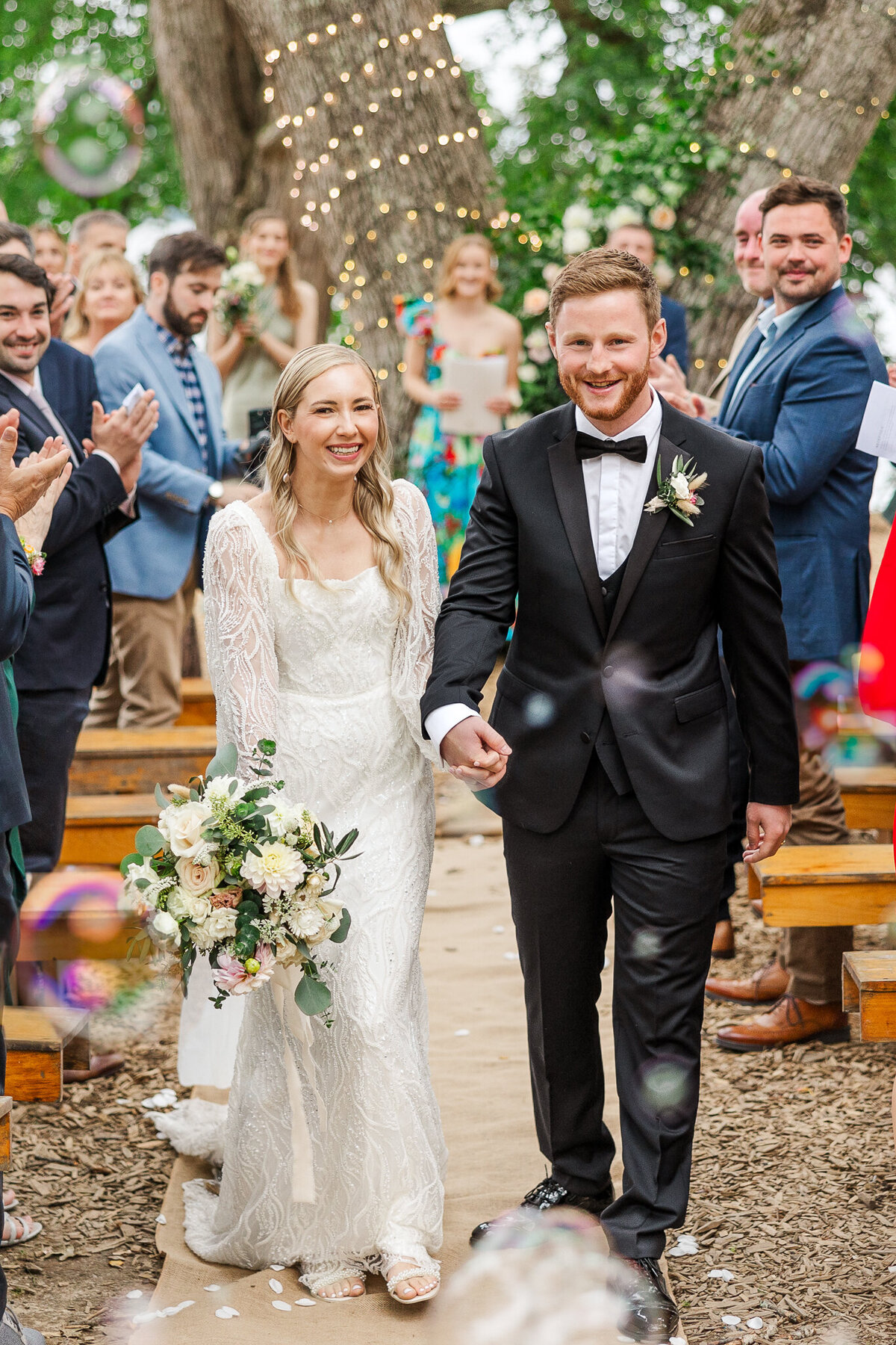 Bride and groom walk down aisle holding hands and smiling as bubbles surround them