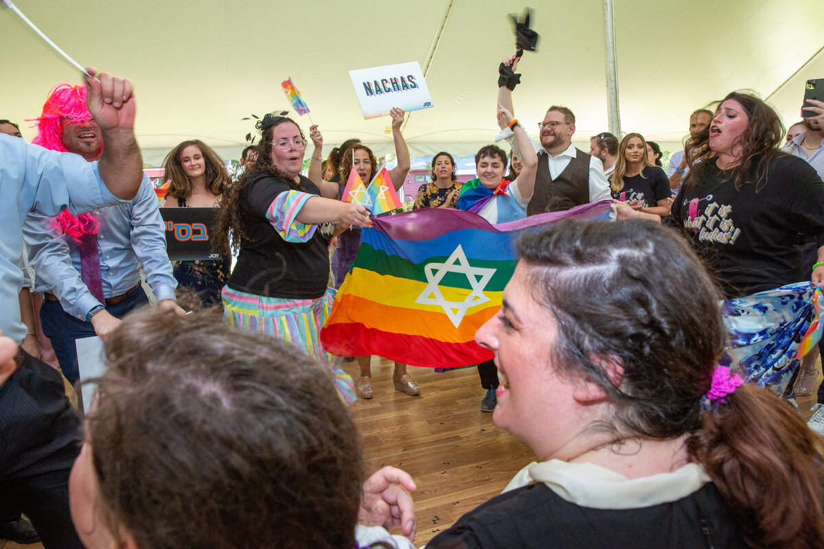 Jewish wedding guests dance with a rainbow flag with Star of David on it for two lesbian brides during shtick reception New York wedding photographer Milestone Images