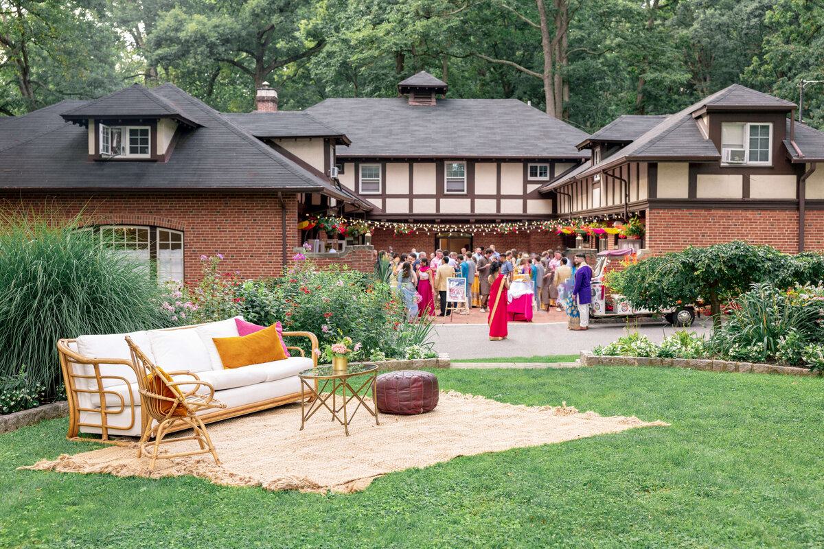 A Sangeet garden party is taking place in front of a charming, rustic house. Guests are gathered under a floral canopy, and a cozy seating area with a white couch and a wicker chair is set up on a rug on the lawn. Lush greenery surrounds the scene.