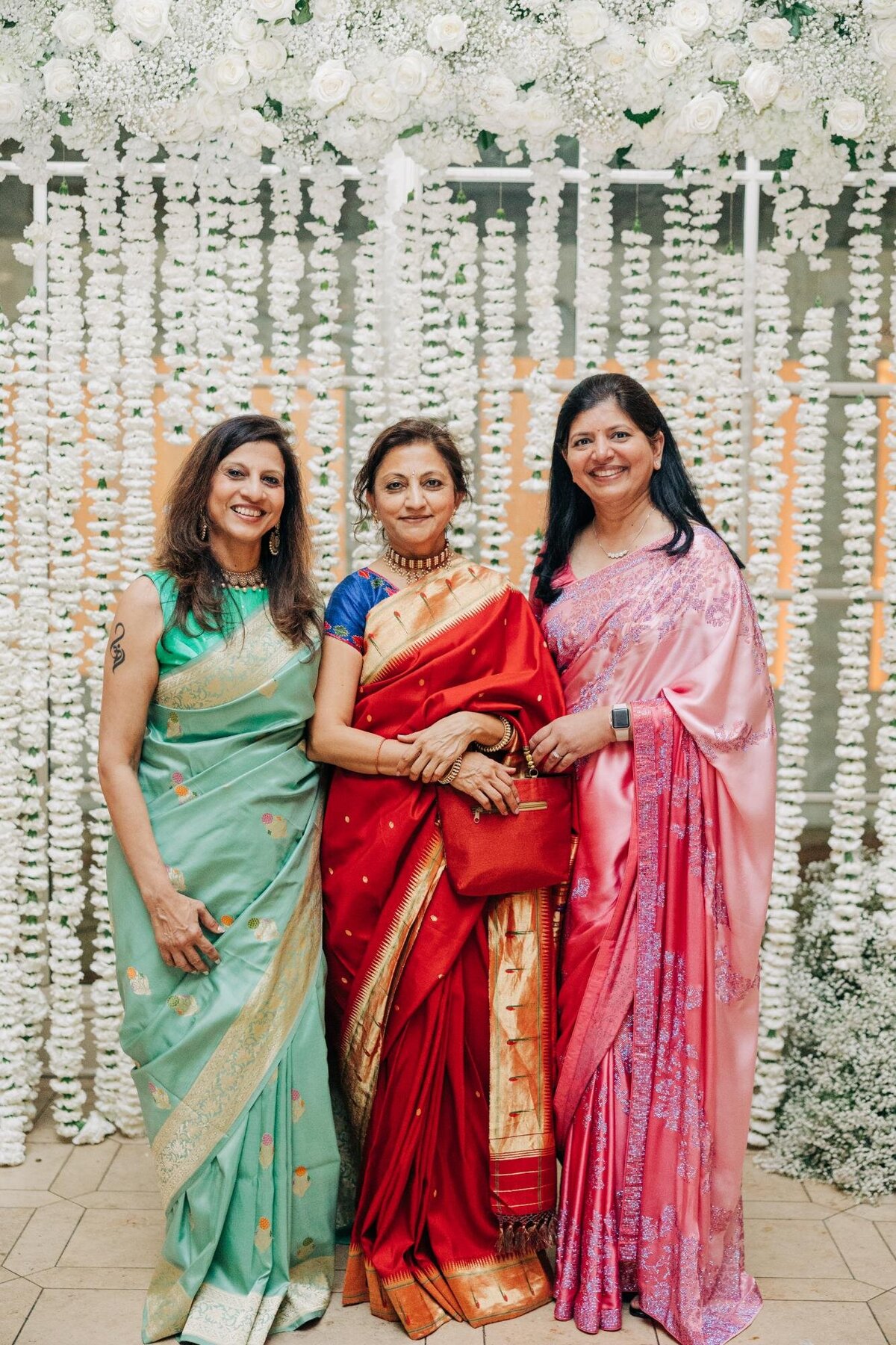 Three women in traditional indian sarees, standing together and smiling at a wedding, with a floral backdrop.