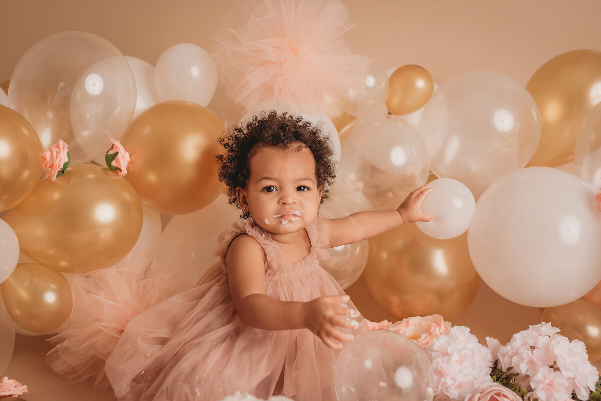 One year old baby girl in pink tulle dress on tan background with neutral colored balloon garland eating her white iced birthday cake with pink roses on it