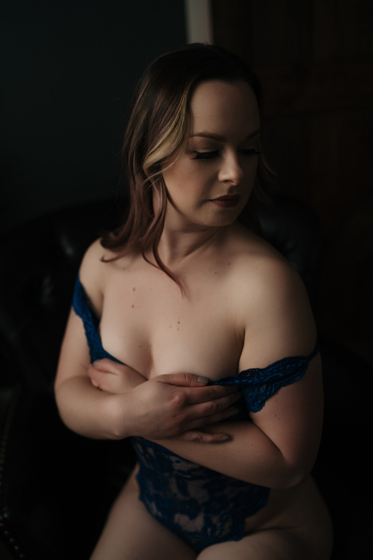 A woman in blue lace lingerie sits in a studio as her straps come off her shoulders in a luxury studio posed by a photographer
